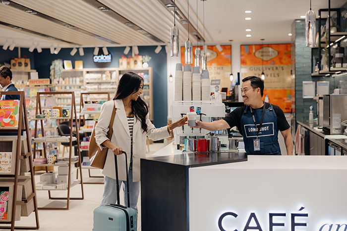 A smiling barista hands a coffee to a traveller with a suitcase at a stylish café inside a bookstore at Gold Coast Airport. Shelves of books and gifts are visible in the background.