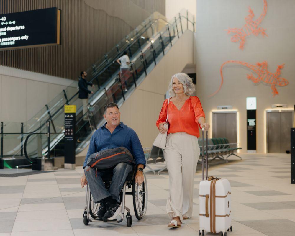 A man in a wheelchair and a woman with a suitcase walk together through a spacious airport terminal. Behind them are escalators, lifts, seating, and a red gecko wall mural, contributing to a bright, modern atmosphere.