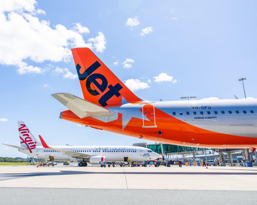A Jetstar plane and two Virgin Australia aircraft are parked at the bay, while another Virgin Australia plane taxis toward the runway for takeoff. The scene is set against a clear blue sky, highlighting the busy airport activity.