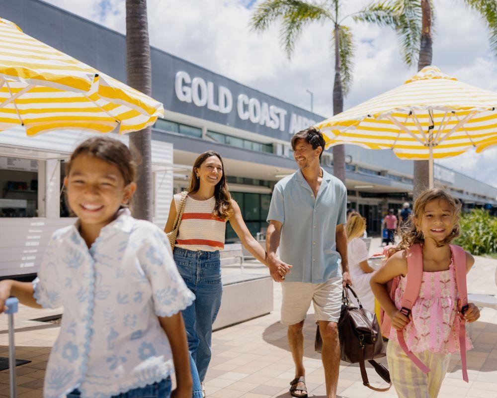 "A smiling family of four walks outside Gold Coast Airport on a sunny day. The parents, a woman in a striped top and jeans and a man in a light blue shirt and shorts, hold hands while carrying travel bags. Two young girls, one wearing a pink backpack and the other pulling a suitcase, walk ahead excitedly. Yellow and white striped umbrellas and palm trees line the walkway, creating a bright and welcoming atmosphere.