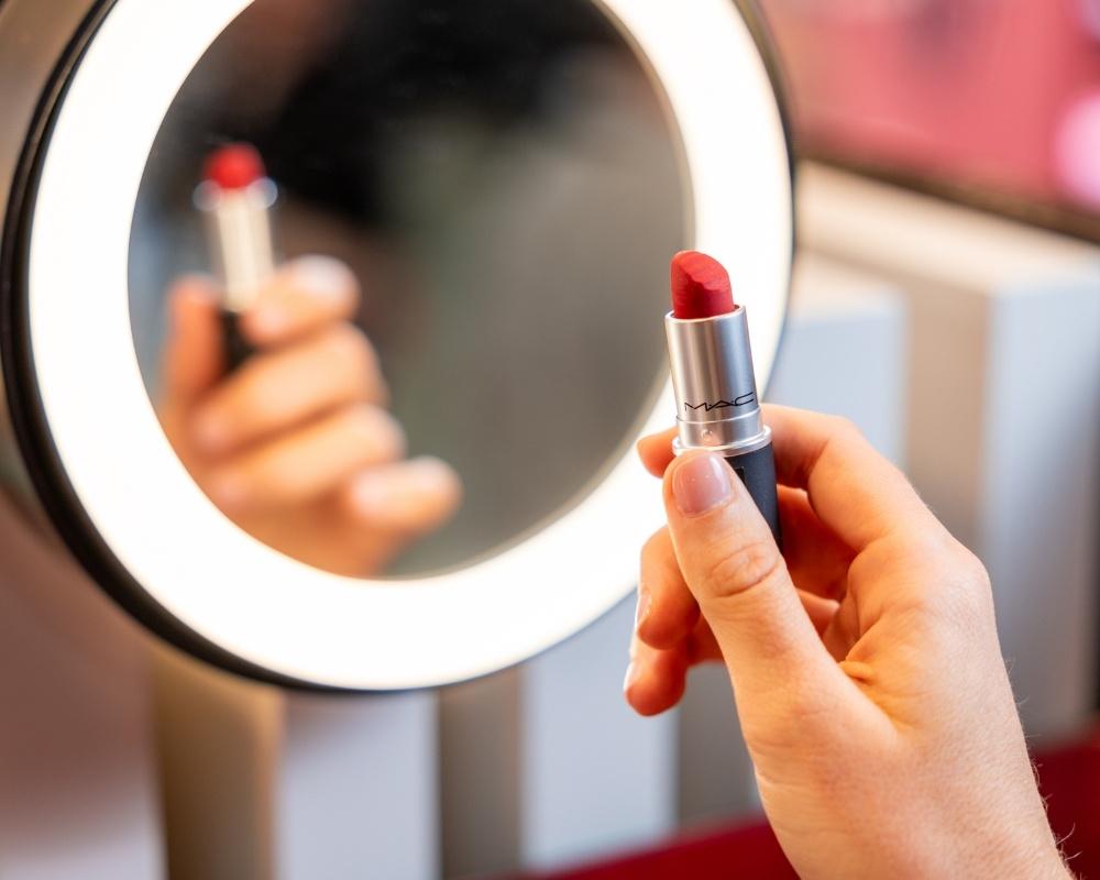 A close-up of a person holding a red lipstick in front of a round, illuminated mirror, with the reflection of the lipstick visible in the mirror.