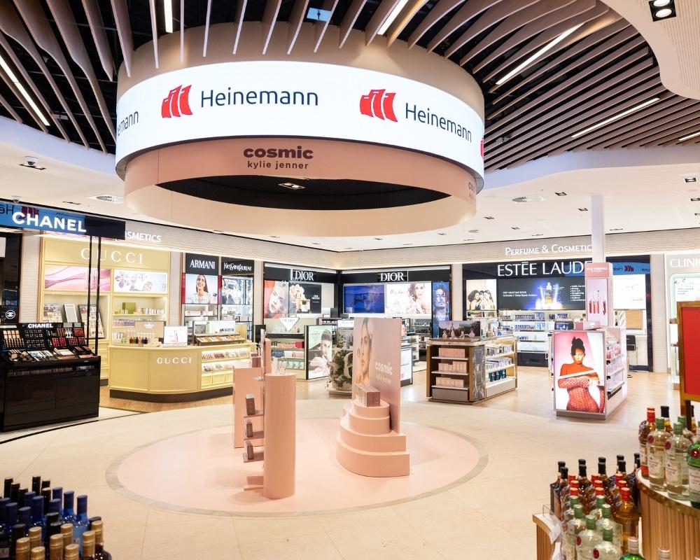The interior of Heinemann Tax and Duty Free at Gold Coast Airport, featuring bright lighting, luxury cosmetics and fragrance displays from brands like Chanel and Estée Lauder, and a central circular display area beneath a large digital sign.
