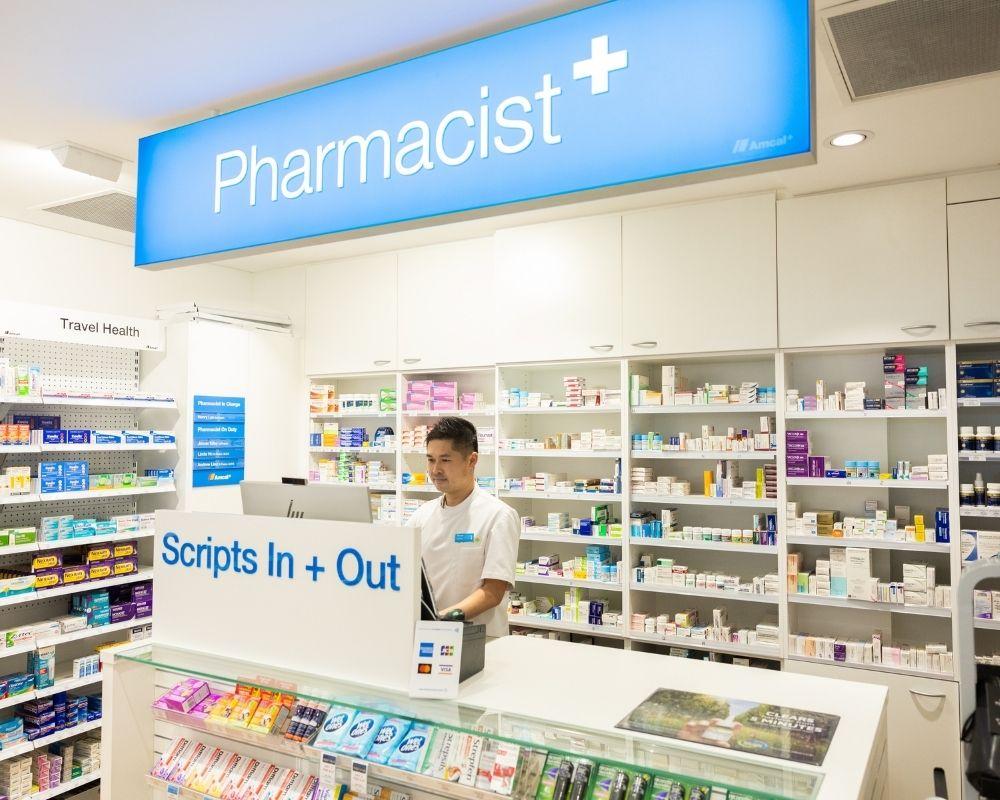 A pharmacist stands behind the counter at a brightly lit pharmacy, surrounded by shelves filled with medications and health products. A blue sign above reads “Pharmacist+” and the counter signage says “Scripts In + Out