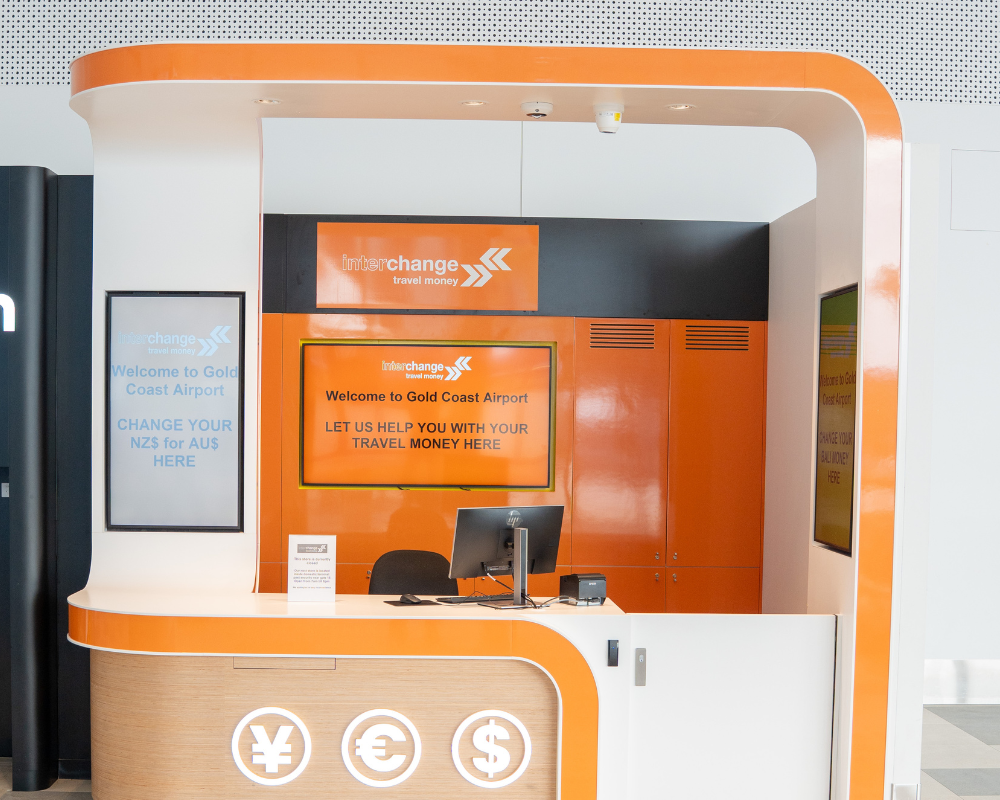Interchange Travel Money currency exchange booth at Gold Coast Airport, with bright orange and white signage displaying messages about exchanging NZ$ for AU$, a counter with a computer, and currency symbols for yen, euro, and dollar.