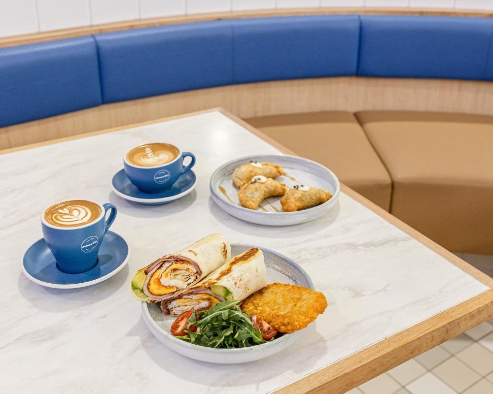 A cosy café setting with a marble table featuring two blue cups of coffee, a breakfast wrap with hash browns and salad, and a plate of pastries. Blue and tan booth seating is visible in the background.