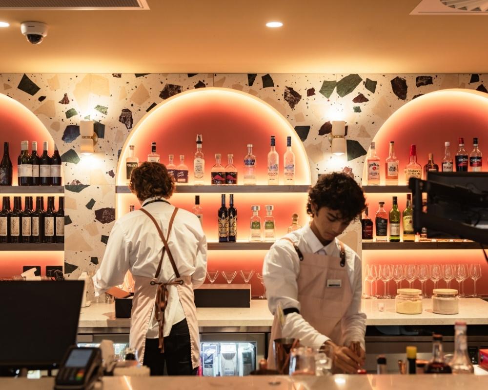 Two bartenders prepare drinks behind a stylish, modern bar with arched backlit shelves displaying bottles of wine and spirits. The wall features terrazzo-style accents and warm pink lighting.