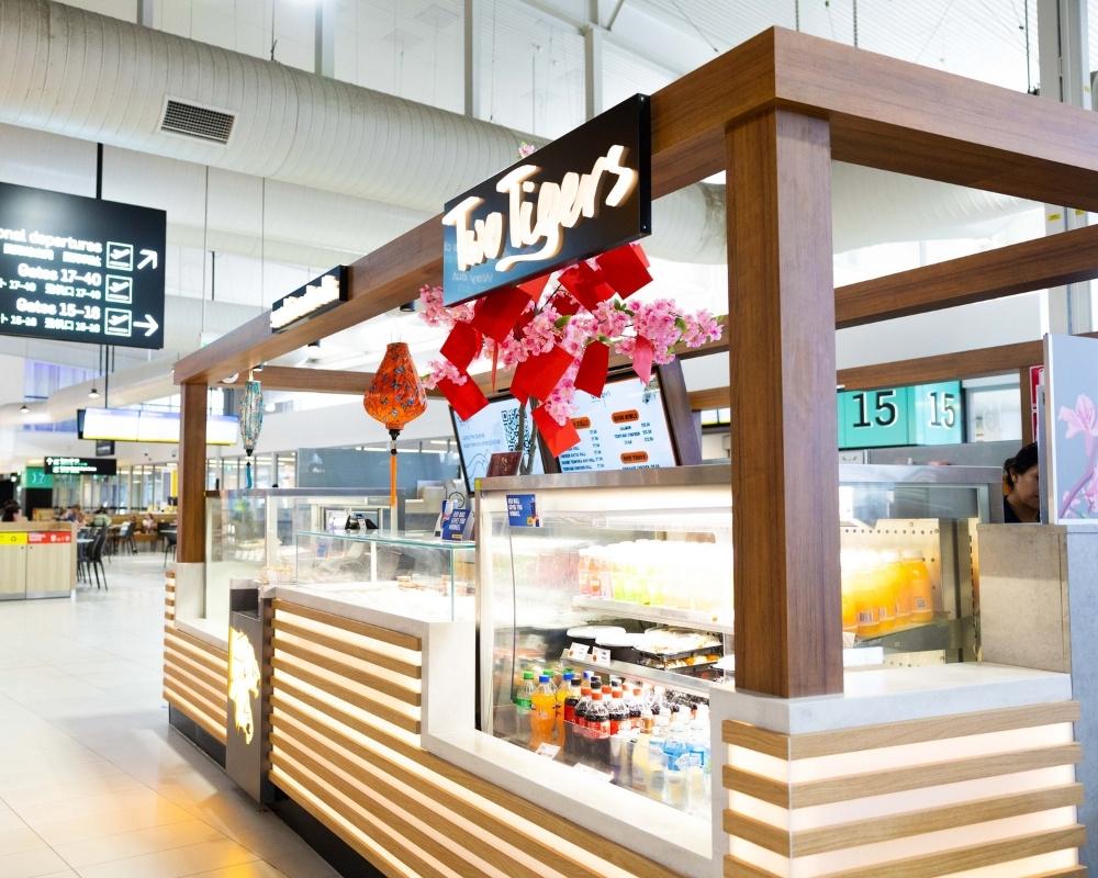 The Two Tigers food kiosk at an airport, featuring a modern wood-accented design with illuminated panels, red lantern decorations, and a fridge stocked with bottled drinks. A departure gate display is visible in the background.