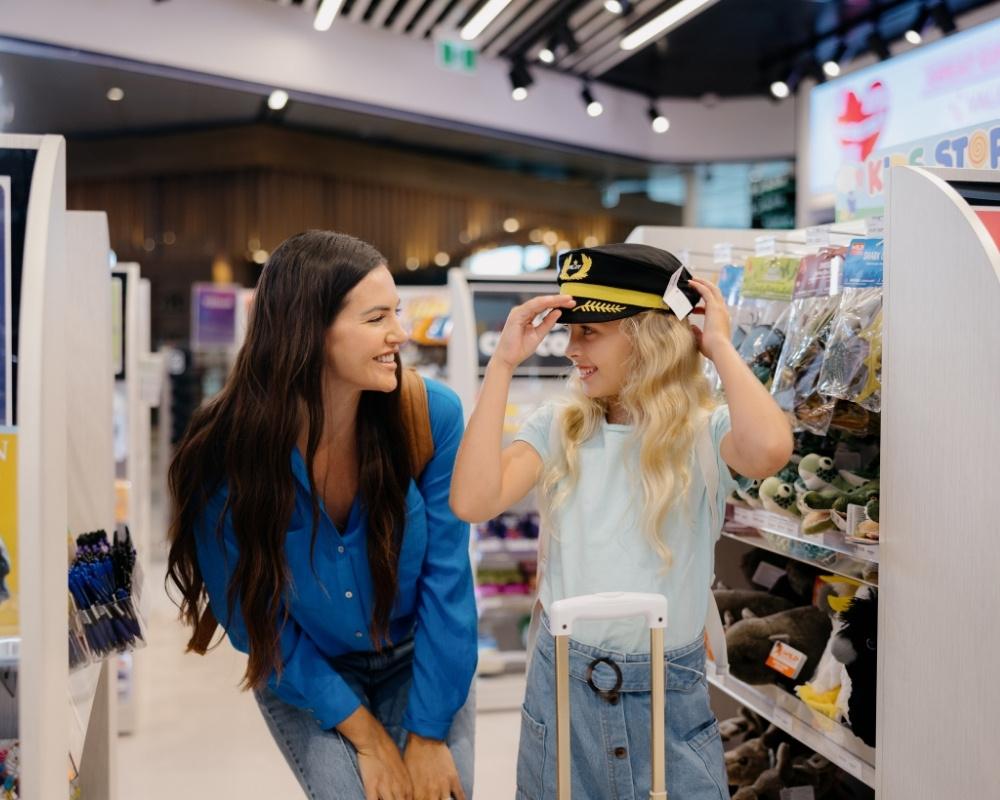 A smiling woman watches a young girl trying on a pilot hat inside a travel retail store. The girl stands beside a white suitcase, and travel accessories line the shelves around them.