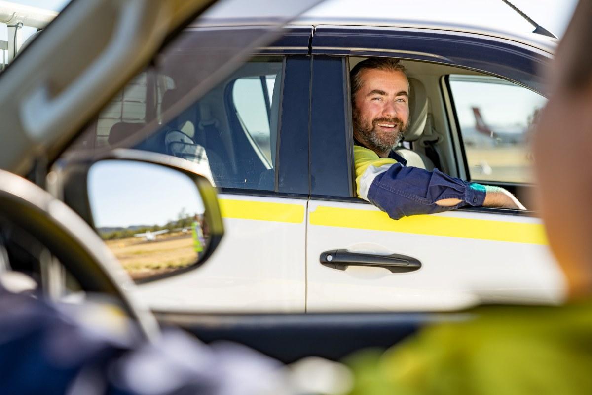 Person seated in the driver's seat of a white vehicle with a yellow stripe, interacting through the open window, airplanes visible in the background at an airfield or airport.