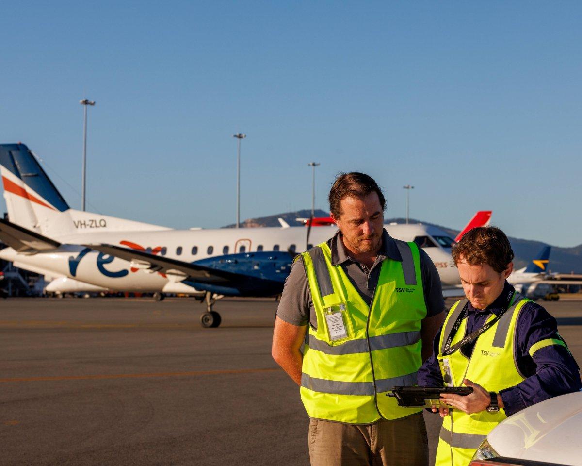 Two airport personnel in high-visibility vests reviewing information on the tarmac near a small aircraft, representing roles requiring ASIC accreditation.