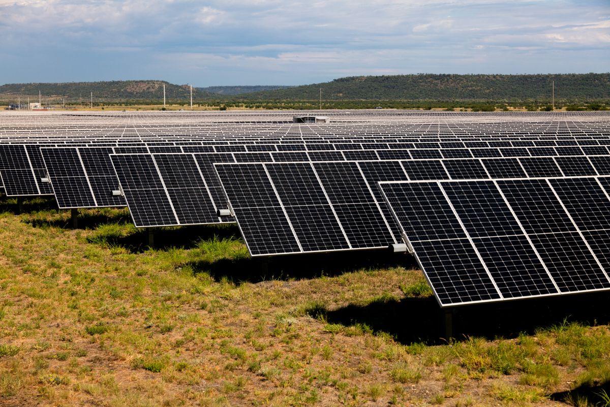 Wide view of a solar panel farm with rows of panels on grassy terrain, set against hills and a partly cloudy sky.
