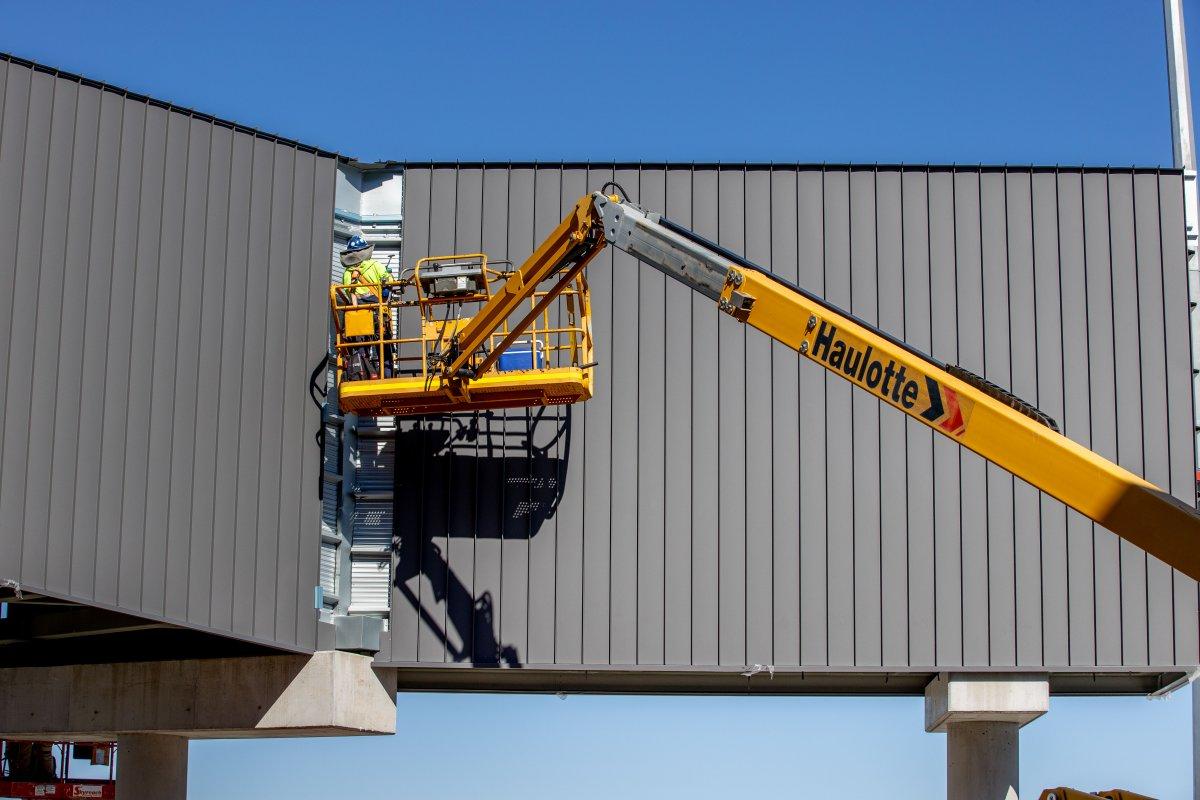 Construction worker operating a yellow boom lift to install metal panels on a building exterior, demonstrating QAL’s commitment to safe and efficient infrastructure development.