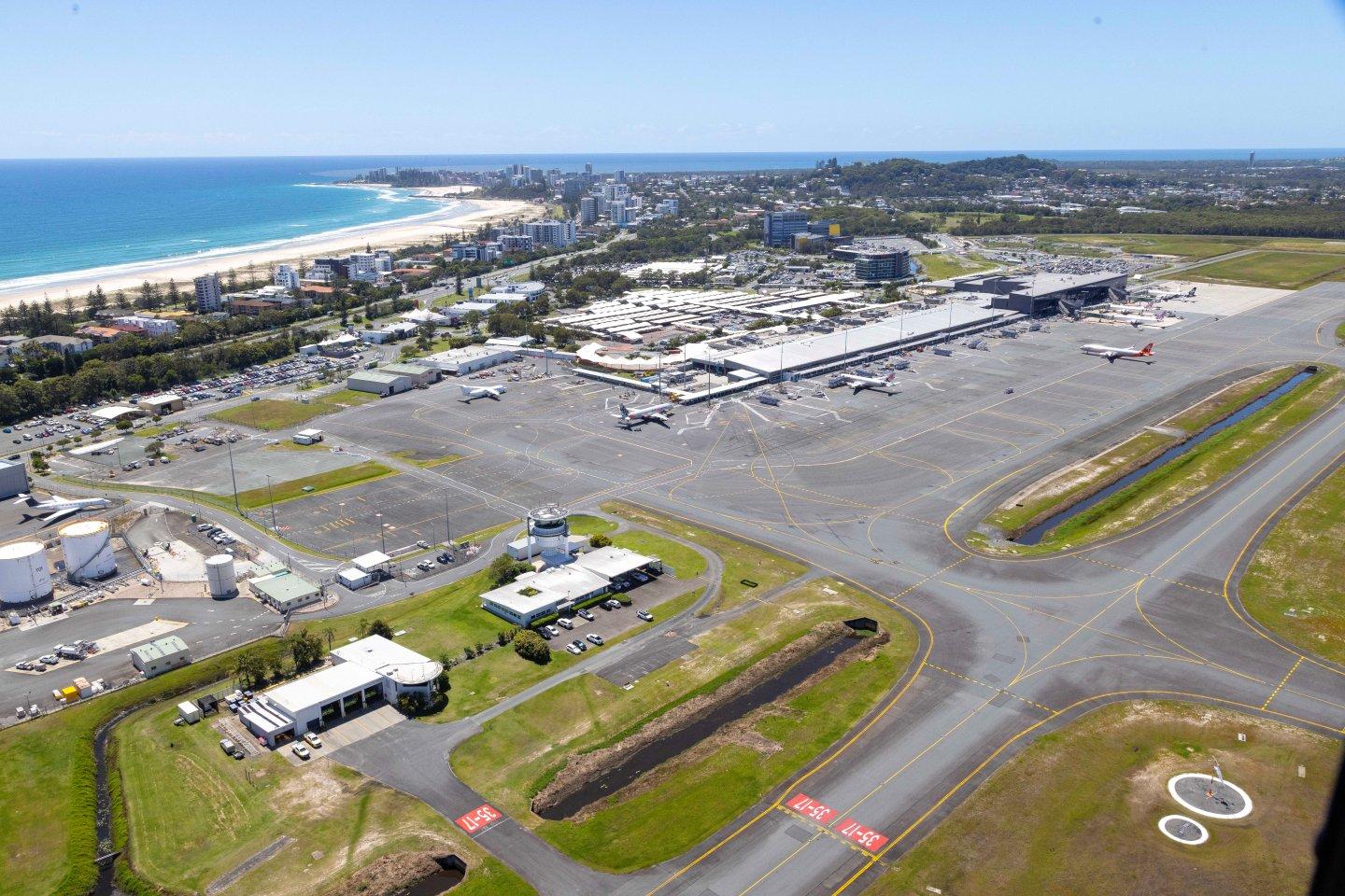 Aerial view of Gold Coast Airport near the coast, showing runways, terminals, and parked aircraft with city and beach in the background.