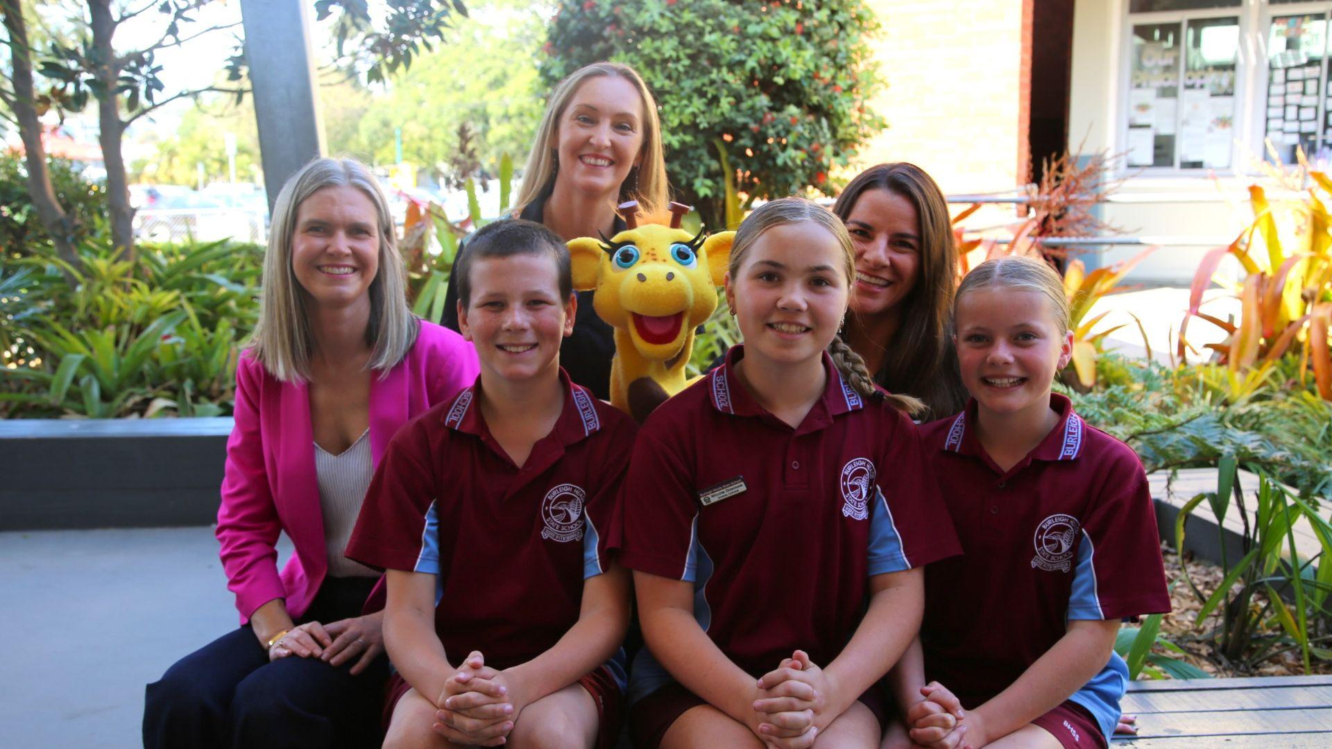 Three adults and three children pose in front of a garden area with various plants. The children wear matching maroon shirts with blue accents and an emblem, while one adult holds a yellow giraffe plush toy. Behind them is a building with windows displaying papers.