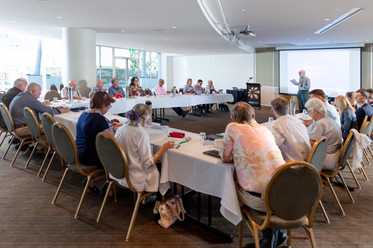 Community consultation meeting held in a conference room with around 20 attendees seated in a U-shaped table arrangement. One person stands at the front near a projector screen, leading the discussion in a formal and professional setting