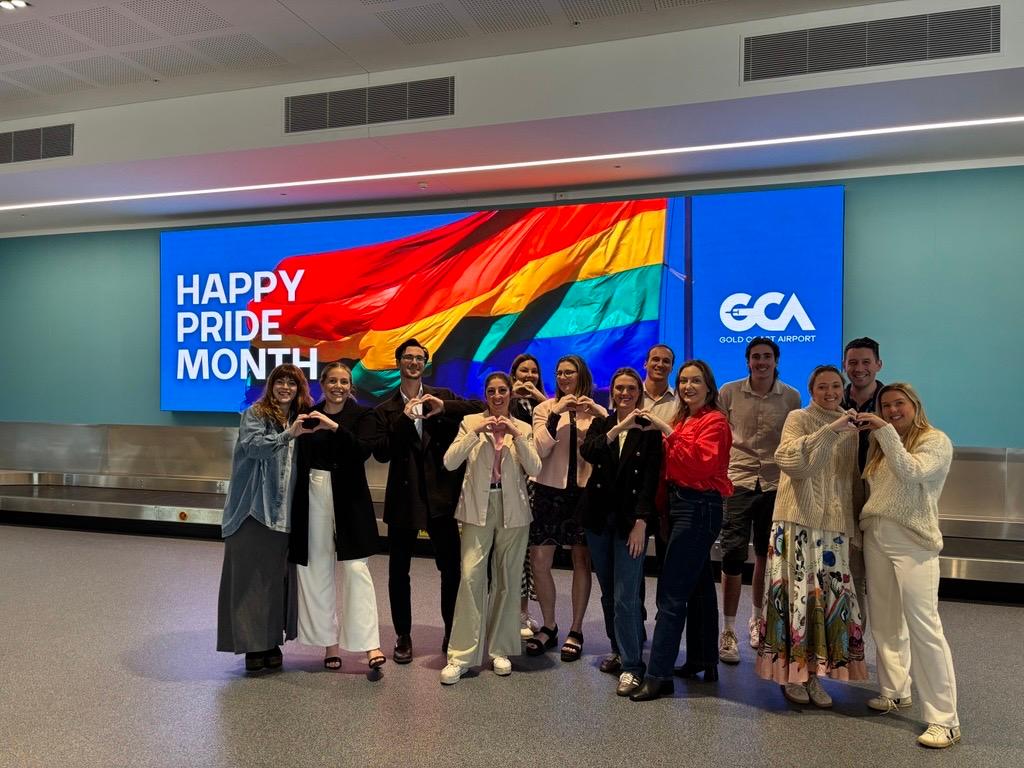 Group of people standing in front of a large digital display at Gold Coast Airport featuring a rainbow flag and the message “Happy Pride Month.” The group poses in two rows, making heart shapes with their hands to celebrate LGBTQ+ inclusion.