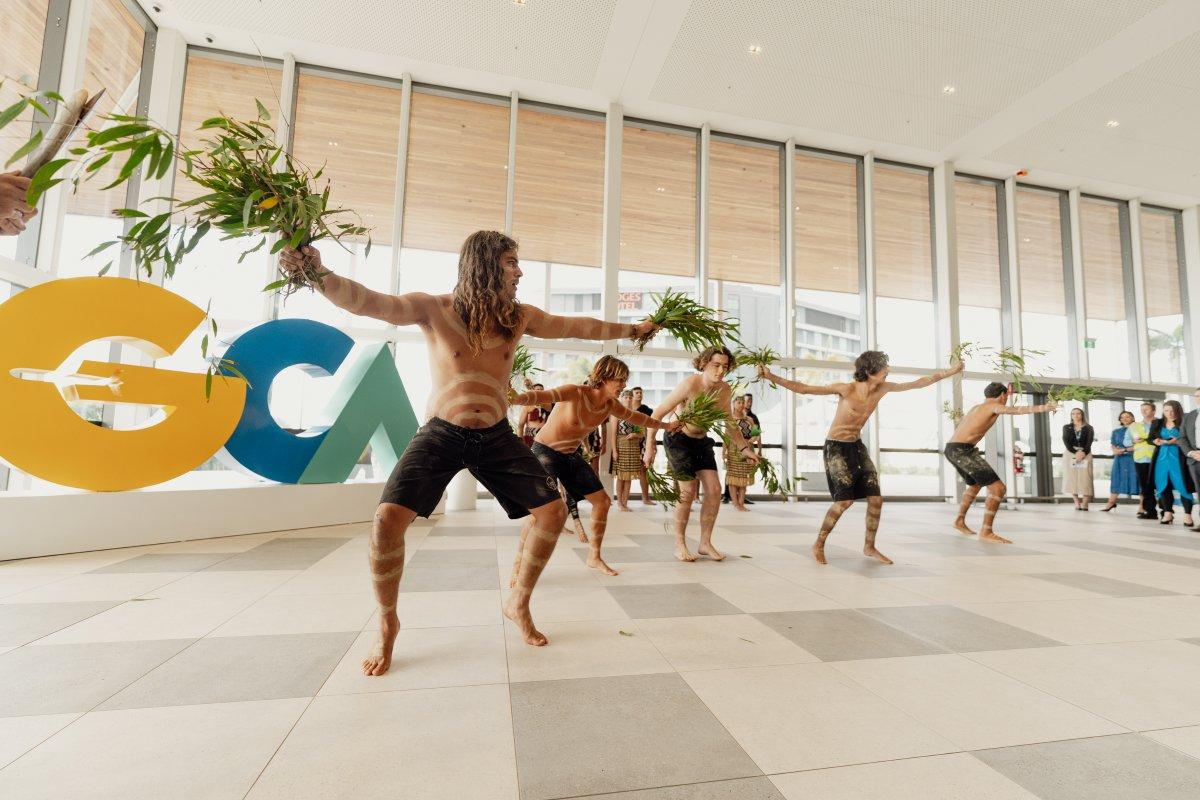 Traditional dance performance inside a GCA's International Arrivals Hall, with dancers holding green foliage and wearing body paint. A large “GCA” sign in yellow, blue, and green is visible in the background, with observers watching the ceremony.