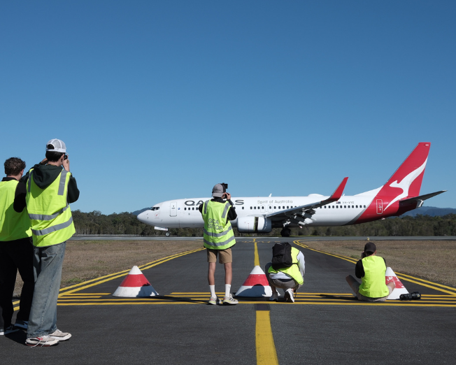 Plane spotters in high-visibility vests photographing a Qantas aircraft on the taxiway under a clear blue sky at Gold Coast Airport.
