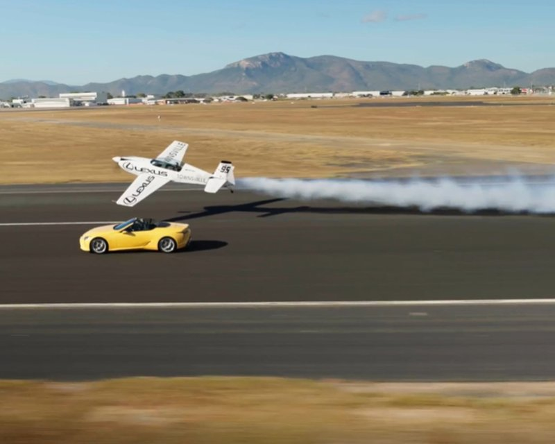Yellow sports car driving on a runway at Townsville Airport with a low-flying white airplane trailing smoke overhead, and airfield buildings and mountains in the background.