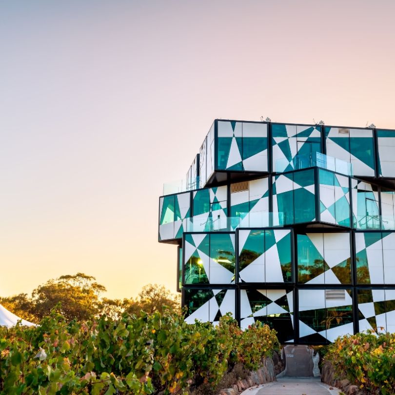 The d’Arenberg Cube in McLaren Vale, South Australia, a striking multi-storey building with an abstract, cube-like design made of glass panels in geometric patterns, surrounded by vineyards at sunset.