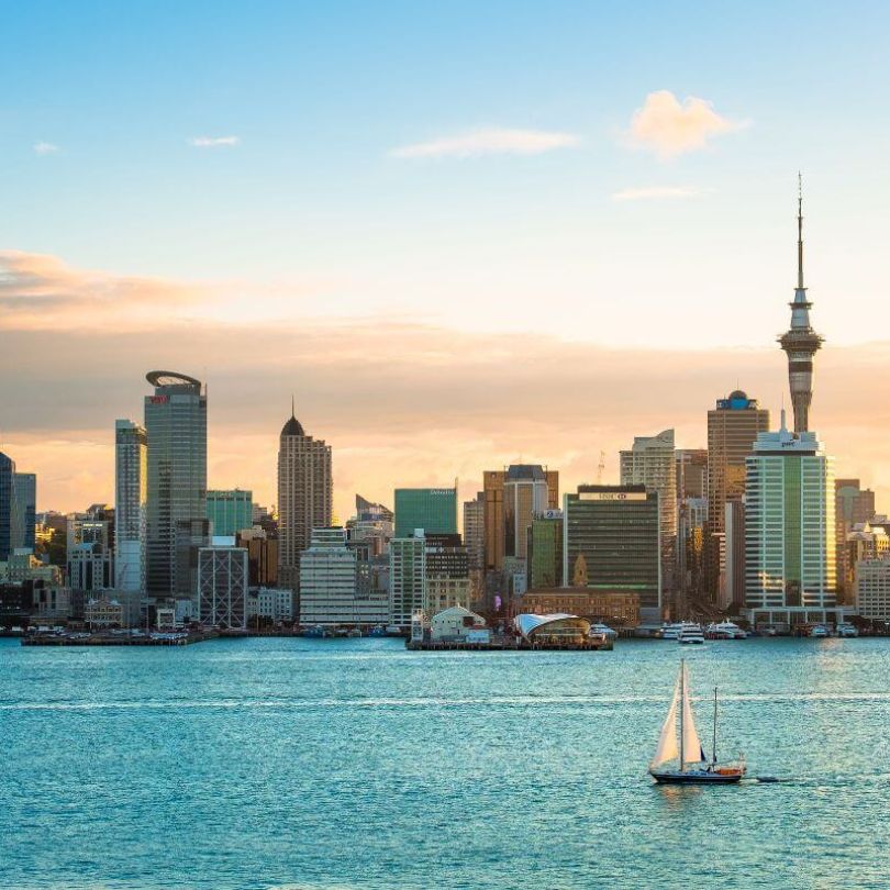 A scenic view of Auckland’s skyline at sunset, featuring the Sky Tower and waterfront buildings. A sailboat glides across the calm harbour waters in the foreground.