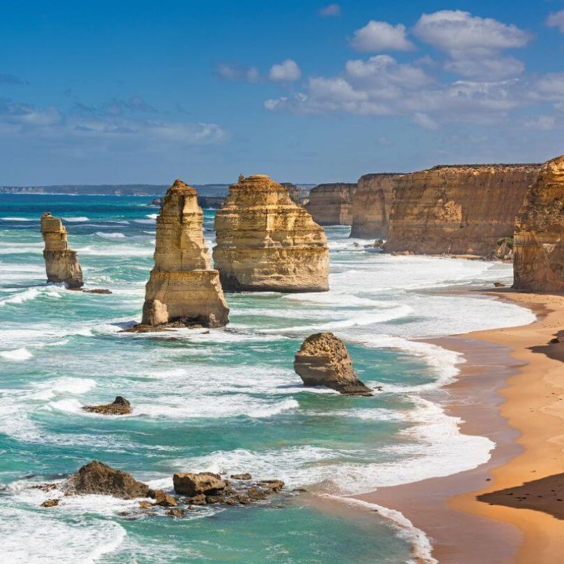 The Twelve Apostles along the Great Ocean Road in Victoria, Australia. Tall limestone stacks rise from the turquoise waters, with waves crashing against the cliffs and a sandy beach stretching along the coastline.