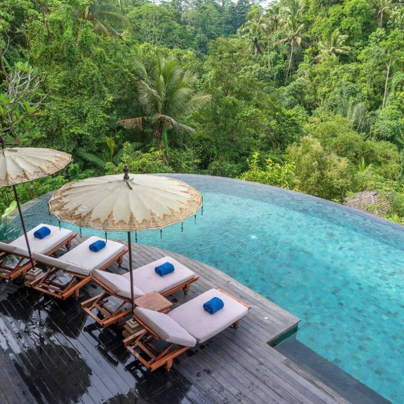 A tranquil infinity pool overlooking lush jungle in Bali, with four sun loungers topped with rolled blue towels and shaded by traditional Balinese umbrellas on a wooden deck.
