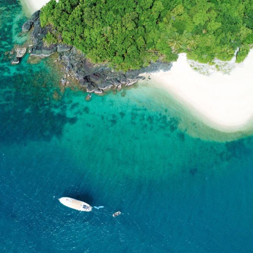  Aerial view of a tropical island with lush green foliage, white sandy beach, and clear turquoise water. A boat floats just offshore, surrounded by vibrant blue ocean and coral reefs.