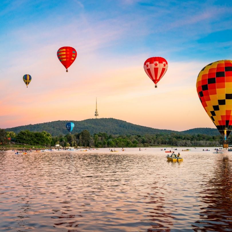 Colourful hot air balloons float above a lake at sunrise in Canberra, Australia. Reflections dance on the water as kayakers paddle below, with Telstra Tower visible atop the hill in the background.