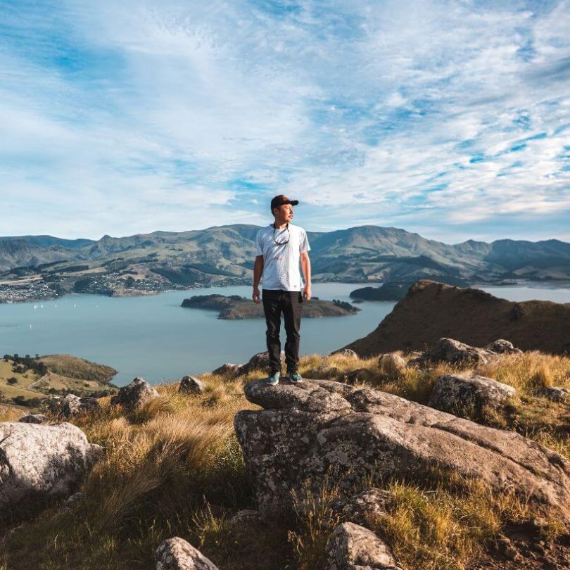  A man stands on a rocky hilltop with panoramic views of Christchurch’s Lyttelton Harbour in New Zealand. Surrounded by golden grass and rugged terrain, he overlooks the deep blue bay and rolling hills under a dynamic sky.