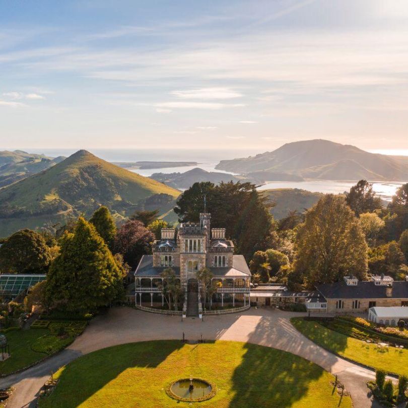 Aerial view of Larnach Castle near Dunedin, New Zealand, surrounded by lush gardens and rolling green hills. In the distance, Otago Harbour glistens under the soft light of late afternoon.