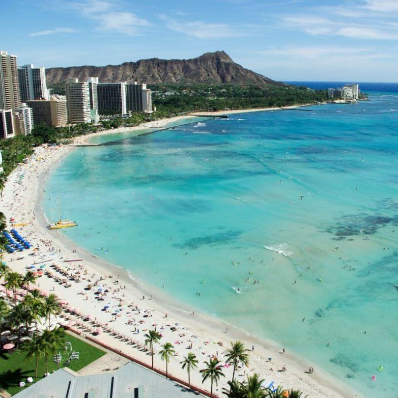 Aerial view of Waikiki Beach in Honolulu, Hawaii, featuring turquoise waters, high-rise hotels, palm trees, and the iconic Diamond Head crater in the background. The beach is lined with sunbathers and swimmers enjoying the tropical coastline.