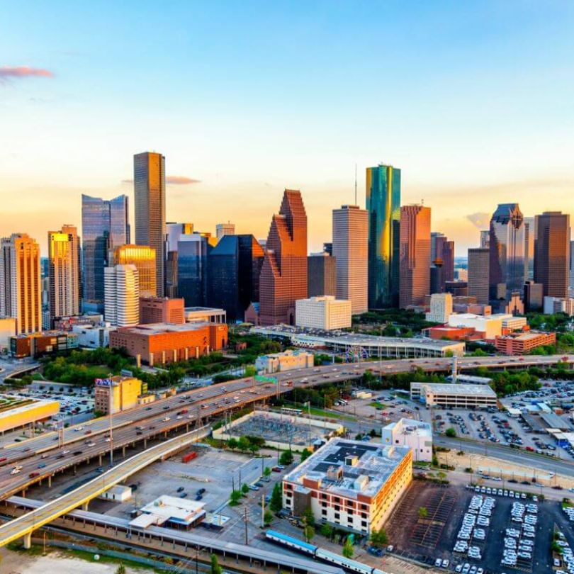 Aerial view of downtown Houston, Texas, at sunset. The skyline features a mix of modern skyscrapers and office towers, with elevated highways and busy roads weaving through the cityscape.