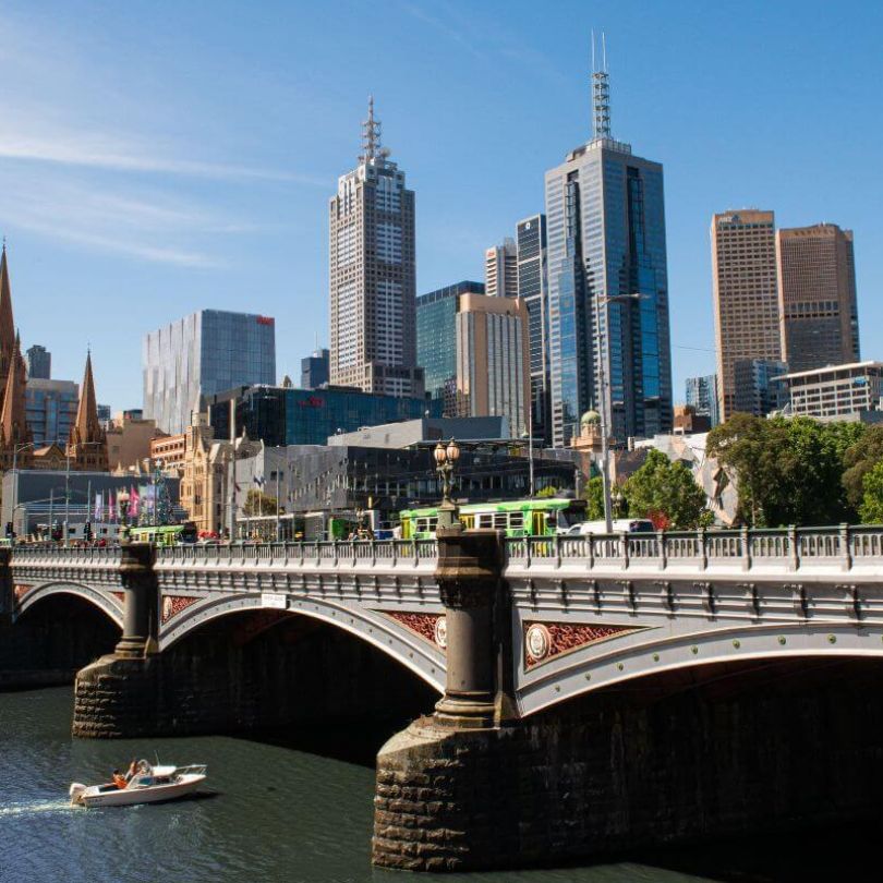Scenic view of Melbourne’s city skyline with a mix of historic and modern buildings, including St Paul’s Cathedral. The Princes Bridge crosses the Yarra River, with a boat cruising beneath and a tram crossing above.