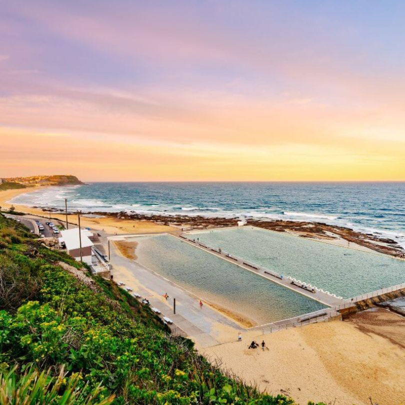 Coastal view of Newcastle Ocean Baths in New South Wales at sunset, with swimmers enjoying the tidal pool beside the sandy beach and waves rolling in from the ocean under a pastel-coloured sky.