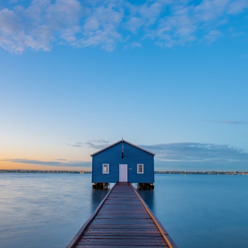 The iconic blue Crawley Edge Boatshed in Perth, Western Australia, sits at the end of a wooden jetty on the calm waters of the Swan River at sunrise, under a clear blue sky.