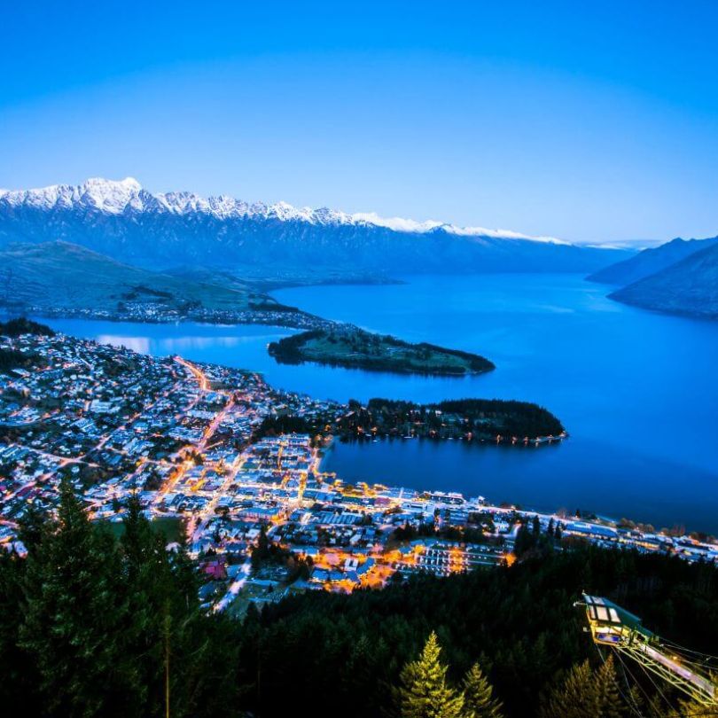  A twilight view of Queenstown, New Zealand, with the town’s lights glowing beside Lake Wakatipu. Snow-capped mountains of The Remarkables range rise in the distance under a deep blue sky.