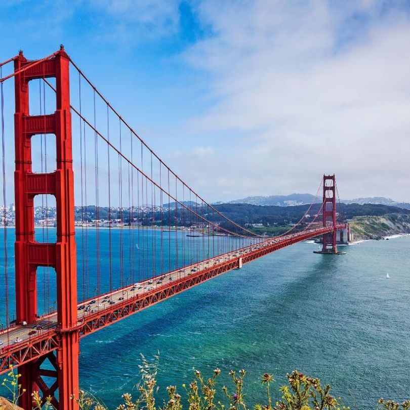 The Golden Gate Bridge in San Francisco, California, stretches across the bay under a partly cloudy sky. The iconic red suspension bridge is surrounded by blue-green waters and rolling hills.