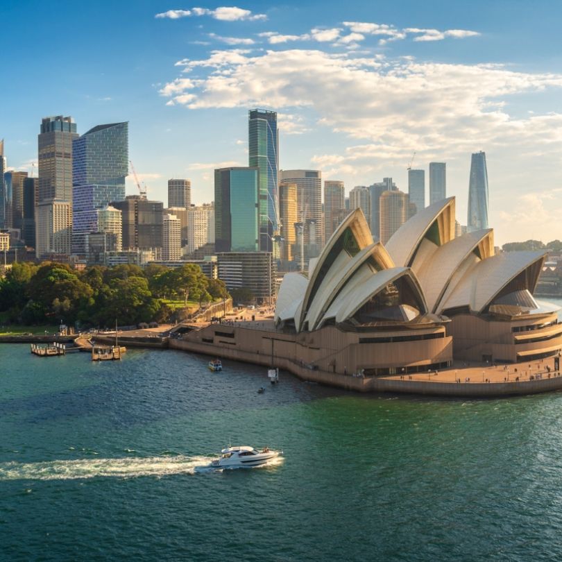 Aerial view of the Sydney Opera House at sunset, surrounded by Sydney Harbour and the city skyline. A white boat cruises across the sparkling water in the foreground.