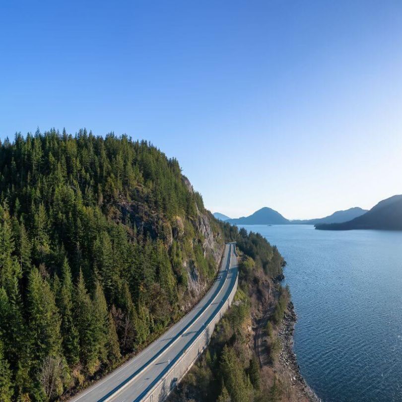 Aerial view of the Sea to Sky Highway near Vancouver, curving between lush green forest and the sparkling waters of Howe Sound, with mountains on the horizon.