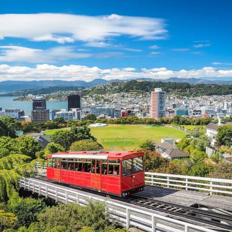 Iconic red Wellington Cable Car travelling uphill with panoramic views over Wellington city, lush gardens, and the harbour on a clear day.