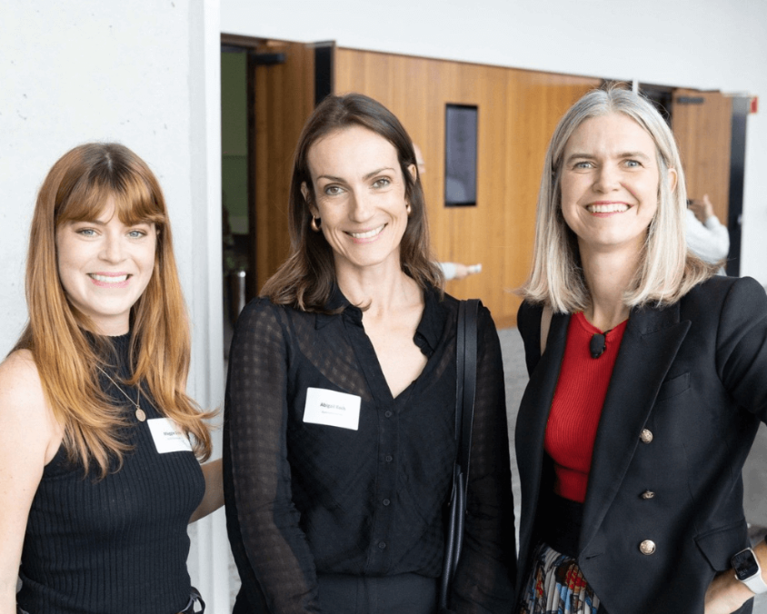 Three women wearing name tags smile at the camera during a professional event, standing in a brightly lit indoor space with timber doors in the background.