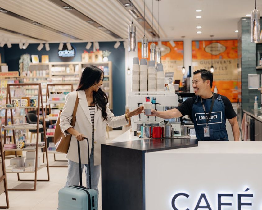 A smiling barista hands a coffee to a traveller with a suitcase at a stylish café inside a bookstore at Gold Coast Airport. Shelves of books and gifts are visible in the background.