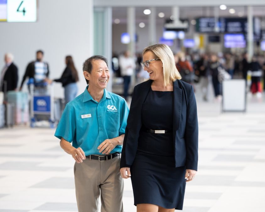 A Gold Coast Airport volunteer and a staff member walk together through a busy terminal, smiling and engaged in conversation, with passengers and flight information screens in the background.