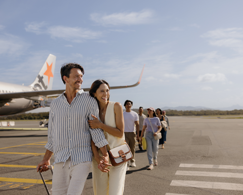 Smiling passengers walk across the tarmac after disembarking a Jetstar aircraft at Gold Coast Airport, with clear skies and mountain views in the distance.