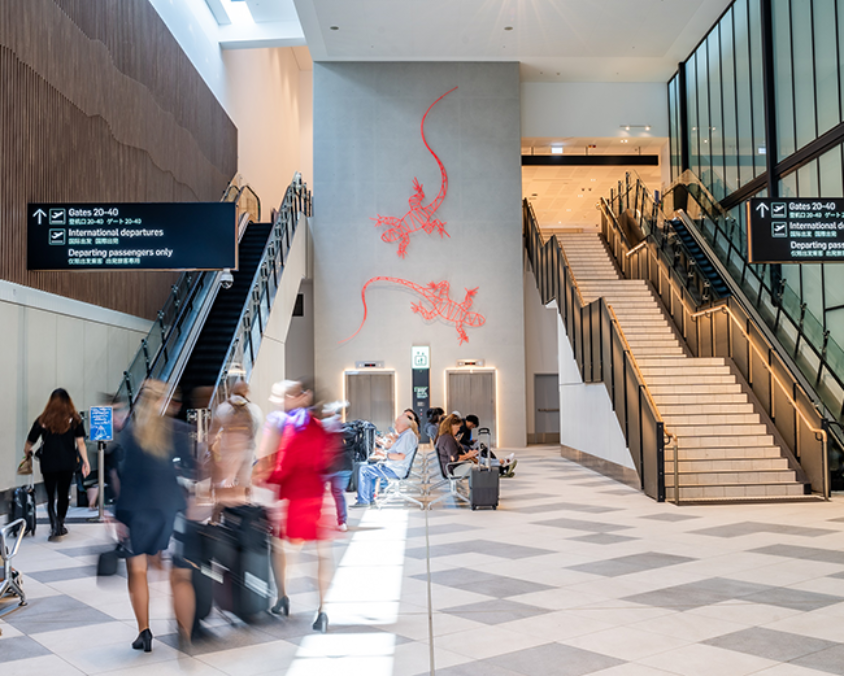 Travellers move through a bright, modern terminal space at Gold Coast Airport, with escalators, stairs, and a wall featuring red gecko artwork. Overhead signage directs passengers to arrivals and baggage claim.