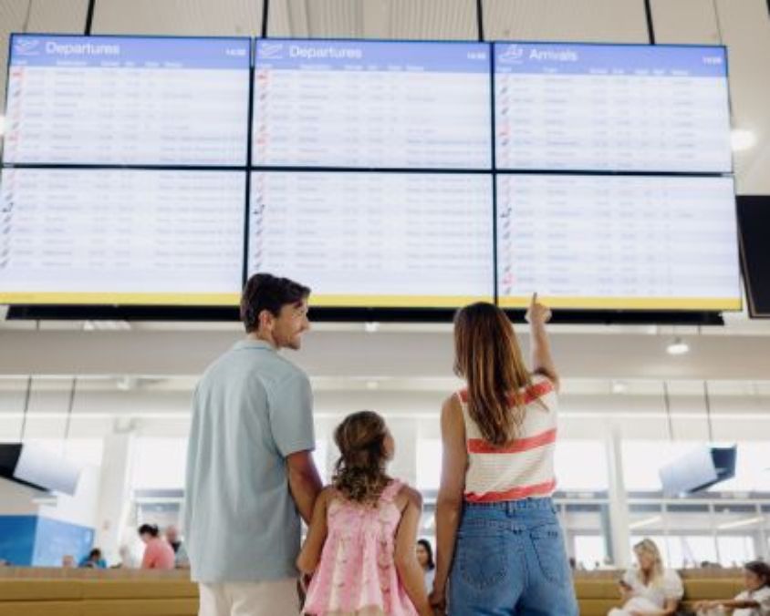 A family of three stands in front of large digital flight information screens at an airport, with the mother pointing toward the departures screen while the father and child look on.
