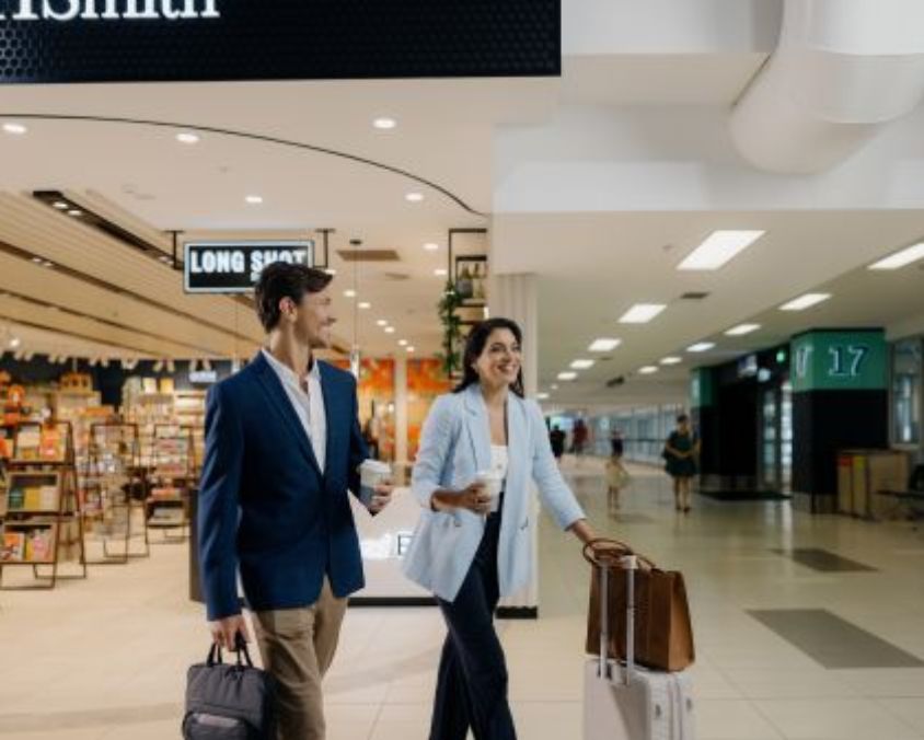 A well-dressed couple walks through the terminal at Gold Coast Airport with luggage and takeaway coffee, passing retail shops and Gate 17 signage in the background.