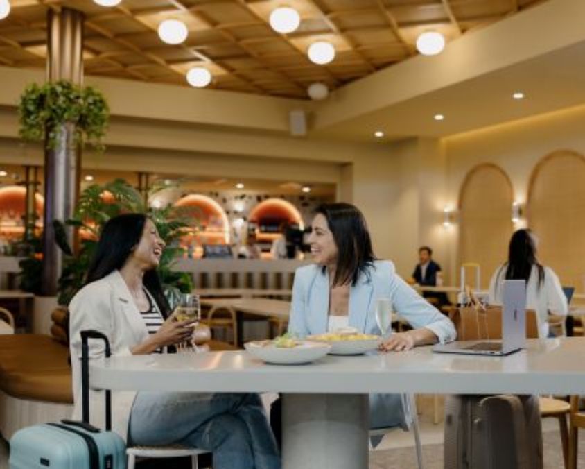 Two women enjoy a meal and conversation at a modern airport dining area, seated at a stylish white table with food and drinks, surrounded by warm lighting and contemporary décor.