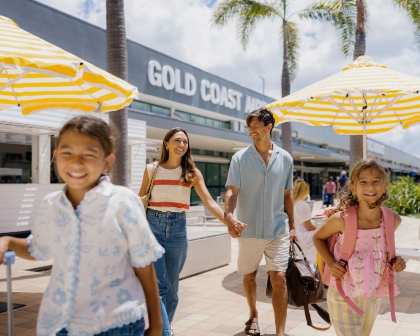 A smiling family of four walks outside Gold Coast Airport on a sunny day. The parents, a woman in a striped top and jeans and a man in a light blue shirt and shorts, hold hands while carrying travel bags. Two young girls, one wearing a pink backpack and the other pulling a suitcase, walk ahead excitedly. Yellow and white striped umbrellas and palm trees line the walkway, creating a bright and welcoming atmosphere.